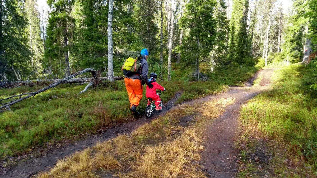 Aikuinen tukee potkupyöräilevää lasta selästä ulkoilureitillä syksyisessä metsässä.