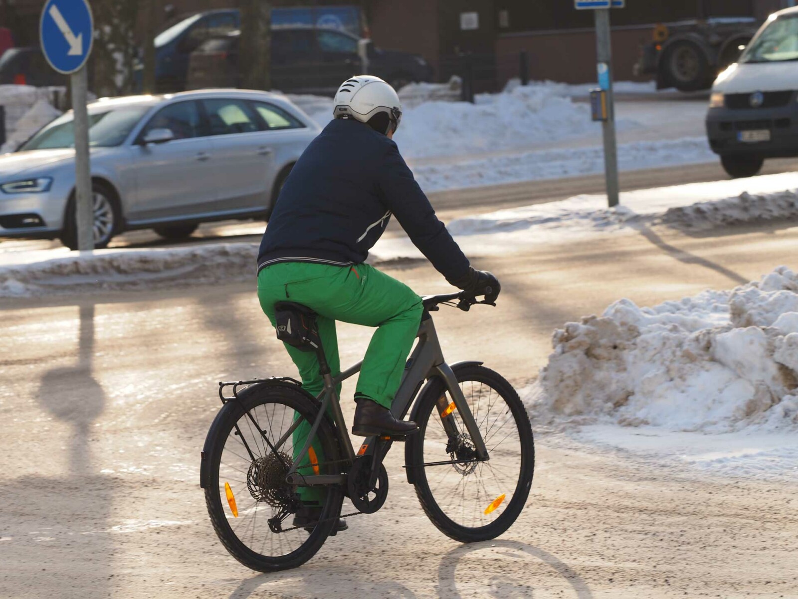 Selkäpuolelta kuvattu vihreisiin housuihin ja siniseen takkiin pukeutunut miesoletettu ajaa sähköpyörällä kaupungin kadulla talvisissa olosuhteissa. Taustalla näkyy autoja ja aurinko paistaa.