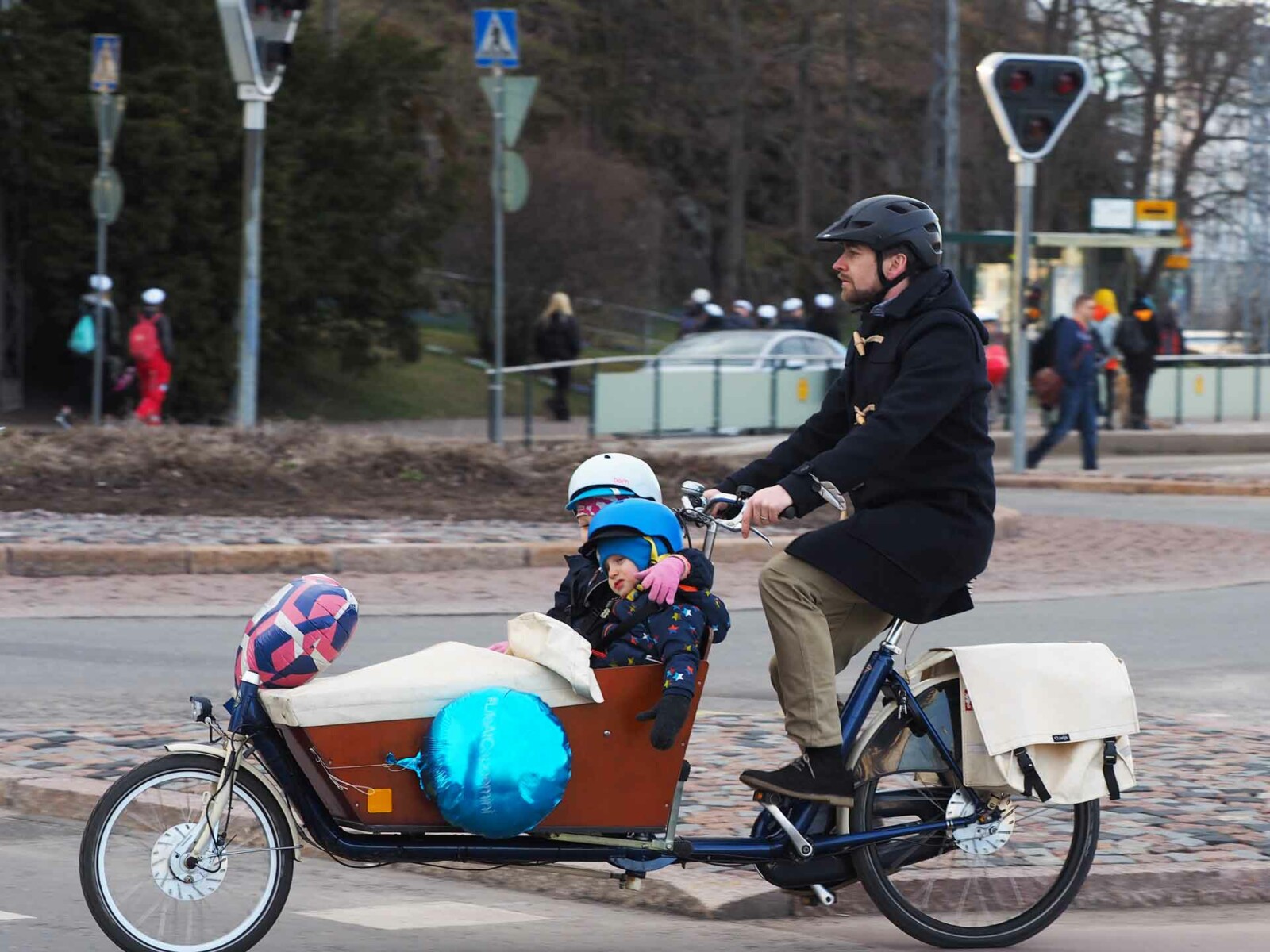 Mies kuljettaa laatikkopyörää kaupungissa kyydissään kaksi lasta.