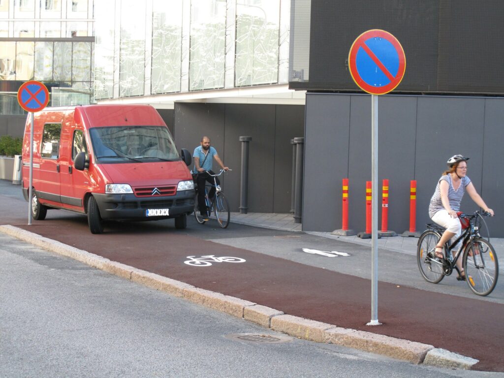 Kuvassa punainen pakettiauto on pysäköinyt keskelle punaisella maalilla merkittyä pyöräkaistaa. Etualalla ajoradan ja pyöräkaistan välissä on pysäköinti kielletty -liikennemerkki sekä pyöräilijä.