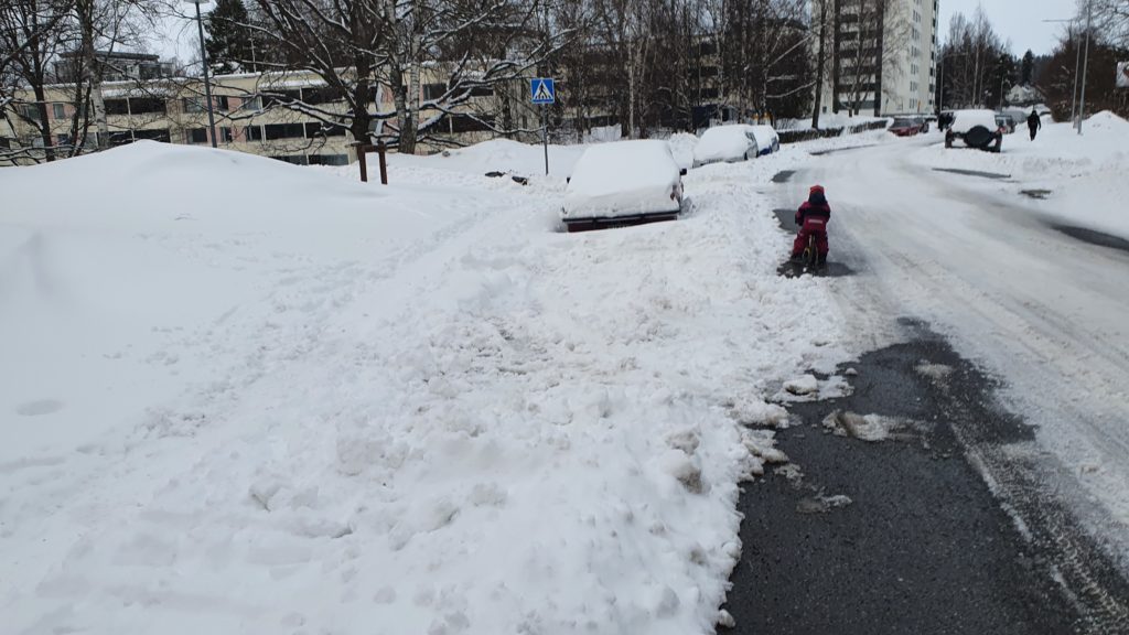 Lapsi potkupyöräilee ajoradalla kun ei voi mennä jalkakäytävälle lumivallien estäessä.